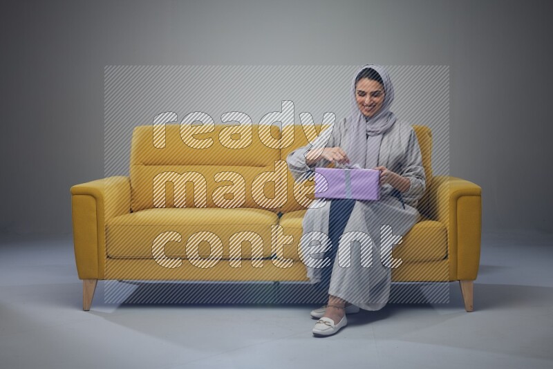 A Saudi woman wearing a light gray Abaya and head scarf sitting on a yellow sofa holding a gift box beside multi color gift boxes eye level on a grey background