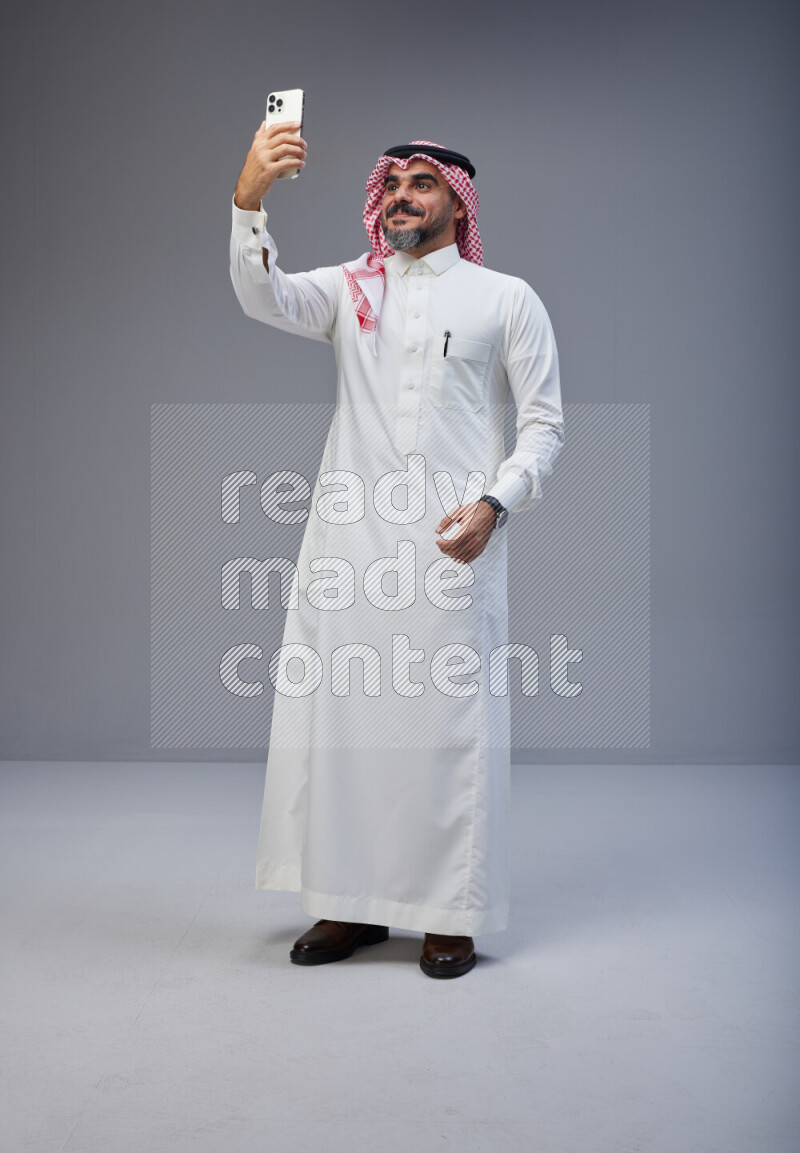 Saudi man Wearing Thob and red Shomag standing taking selfie on Gray background