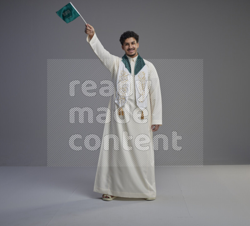 A Saudi man standing wearing thob and saudi flag scarf and holding small saudi flag on gray background