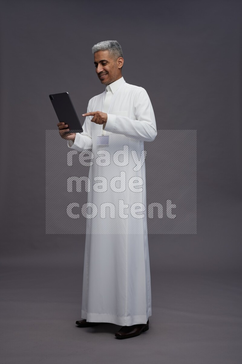 Saudi man wearing thob with neck strap employee badge standing working on tablet on gray background