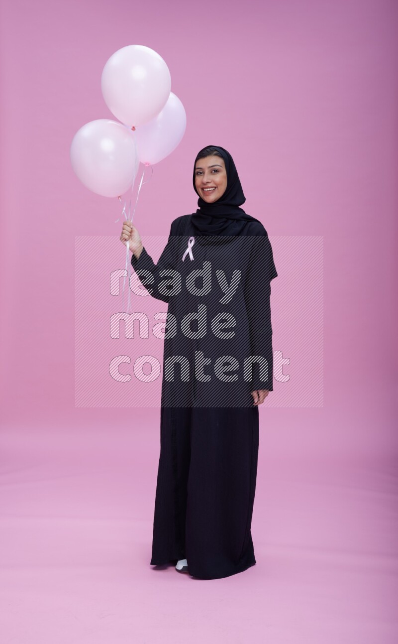 Saudi woman wearing pink ribbon on Abaya standing holding balloons on plain pink background