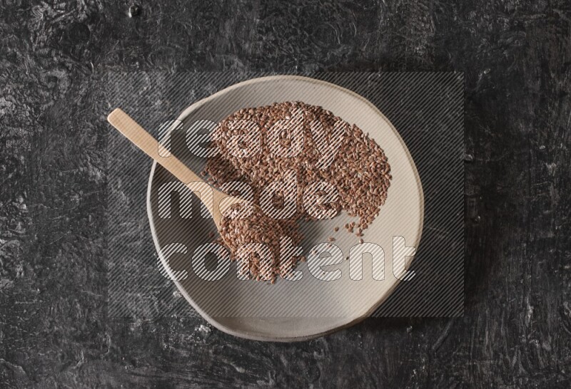 A multicolored pottery plate full of flaxseeds and wooden spoon full of seeds on a textured black flooring