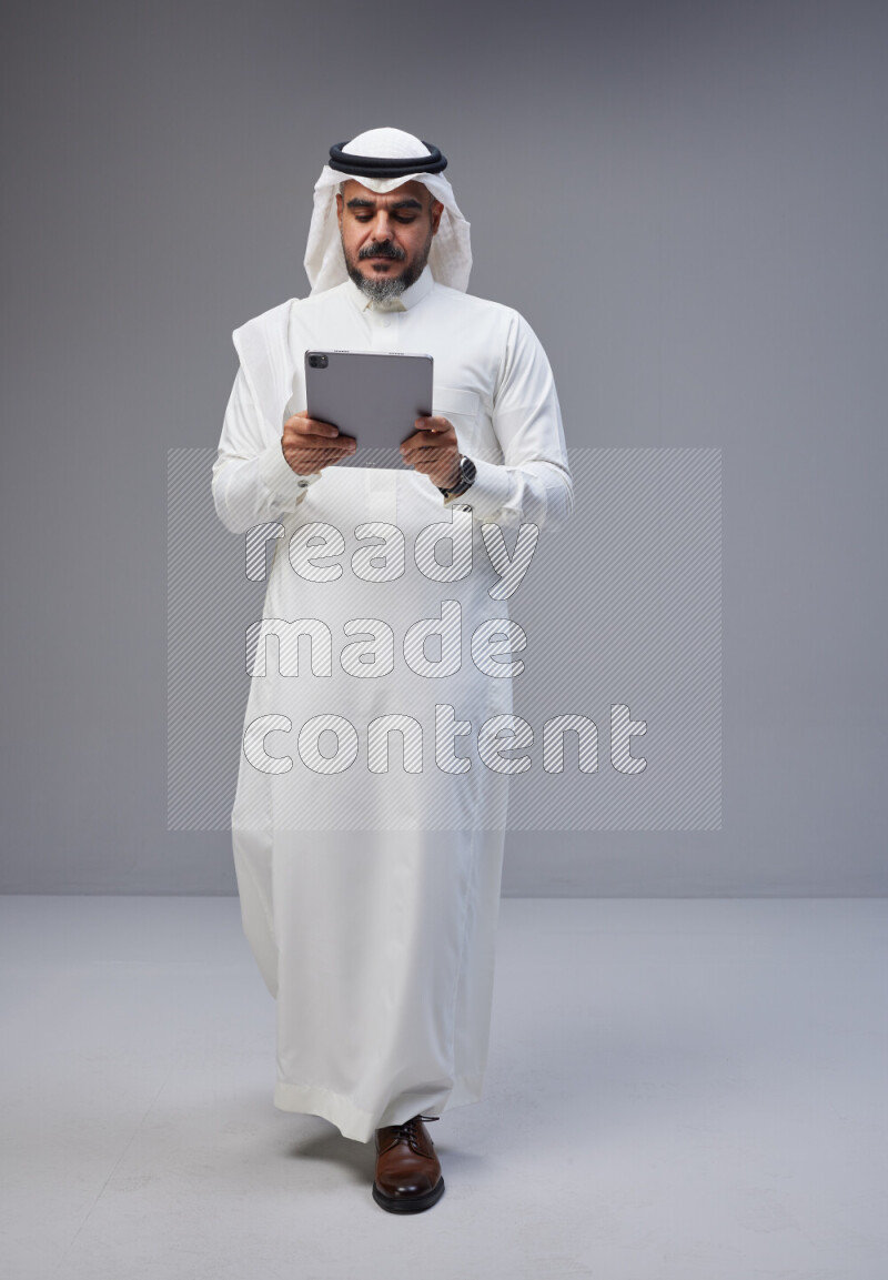 Saudi man Wearing Thob and white Shomag standing working on tablet on Gray background