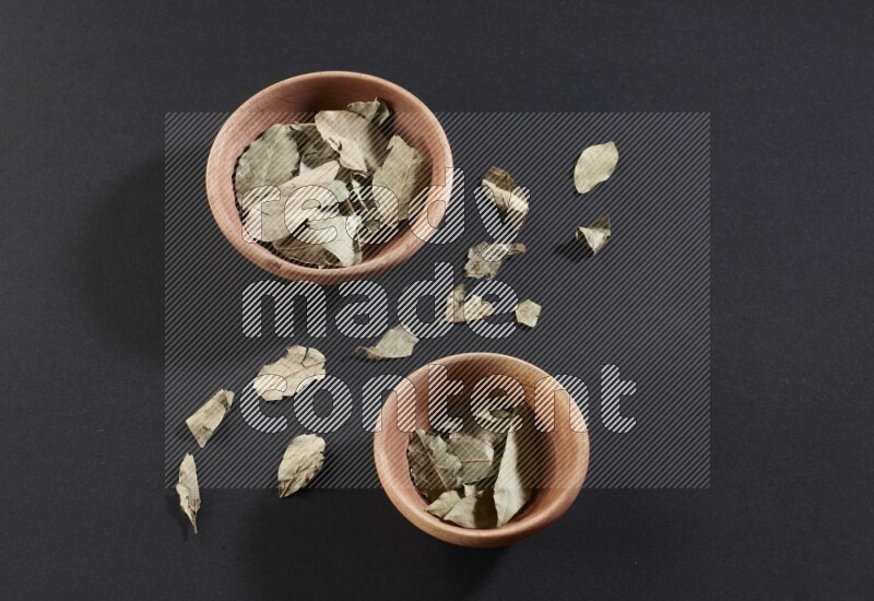 2 wooden bowls full of dried bay leaves with more leaves spread on black flooring