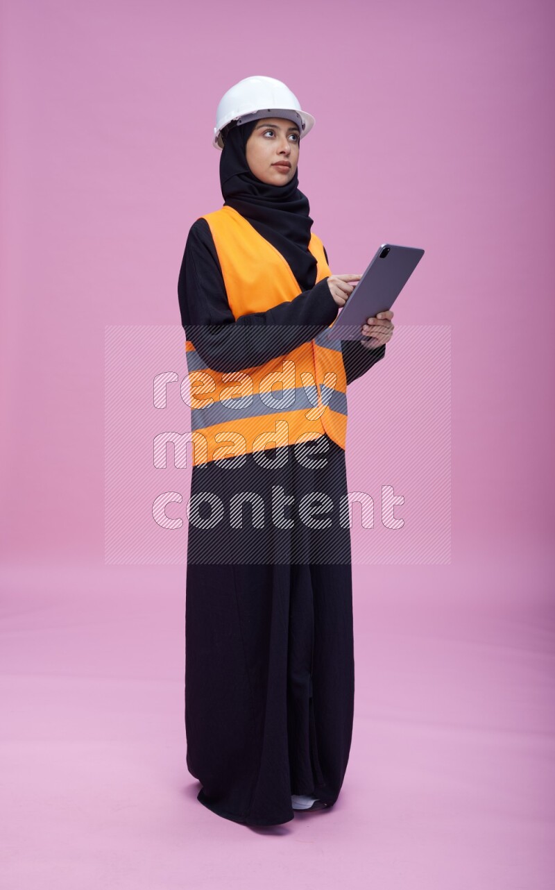 Saudi woman wearing Abaya with engineer vest and helmet standing working on tablet on pink background