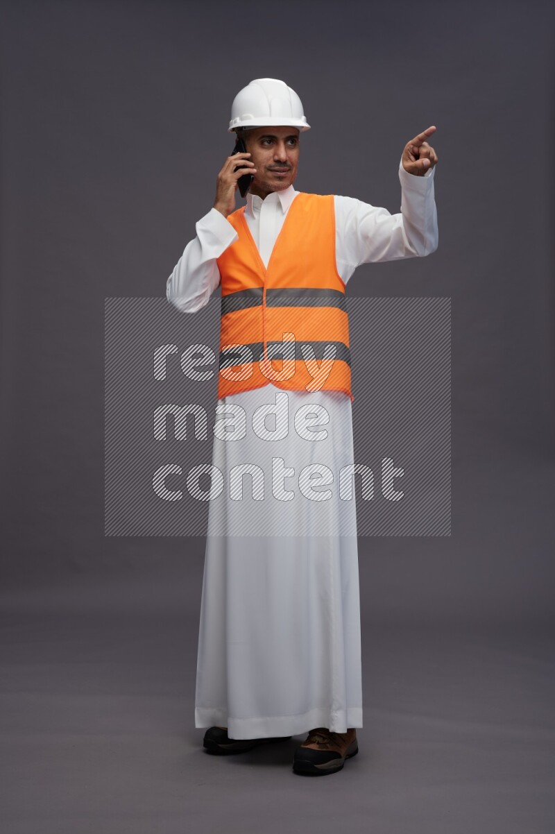 Saudi man wearing thob with engineer vest standing talking on phone on gray background