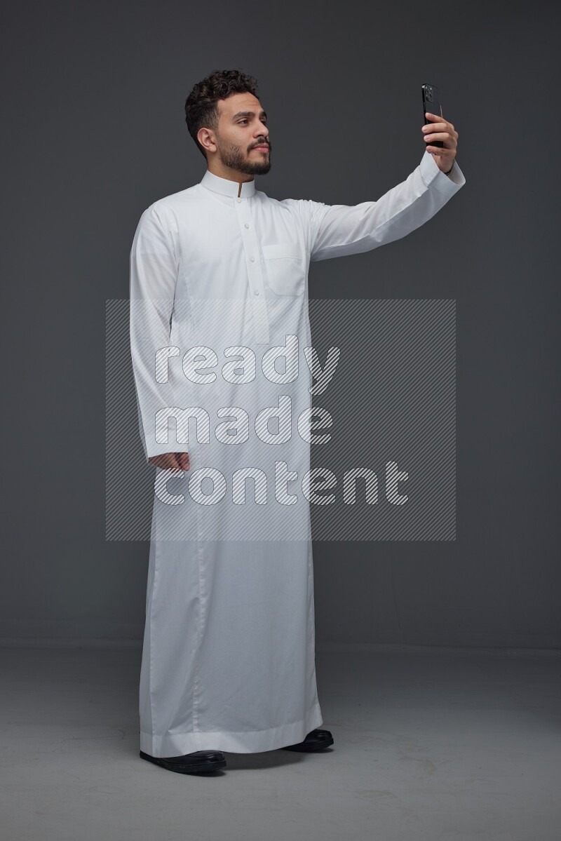 A Saudi man wearing Thobe and taking selfie with his phone making different poses eye level on a gray background
