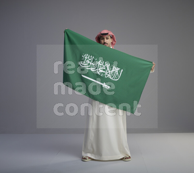 A saudi man standing wearing thob and red shomag holding big saudi flag on gray background