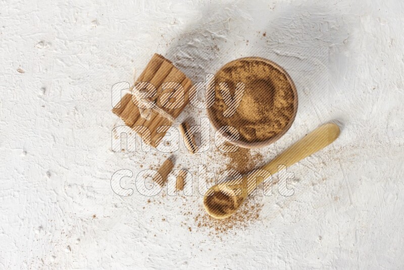 Cinnamon sticks stacked and bounded beside a wooden bowl full of cinnamon powder and a wooden spoon full of powder on white background