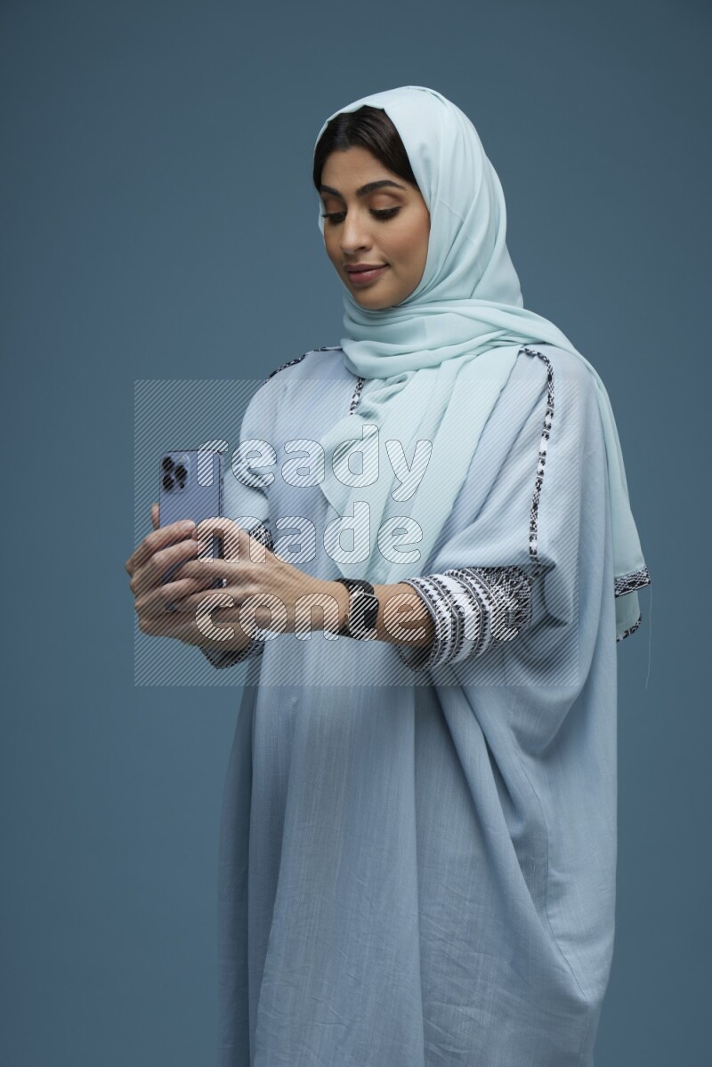 A Saudi woman Taking a Selfie on a blue background wearing a blue Abaya with hijab