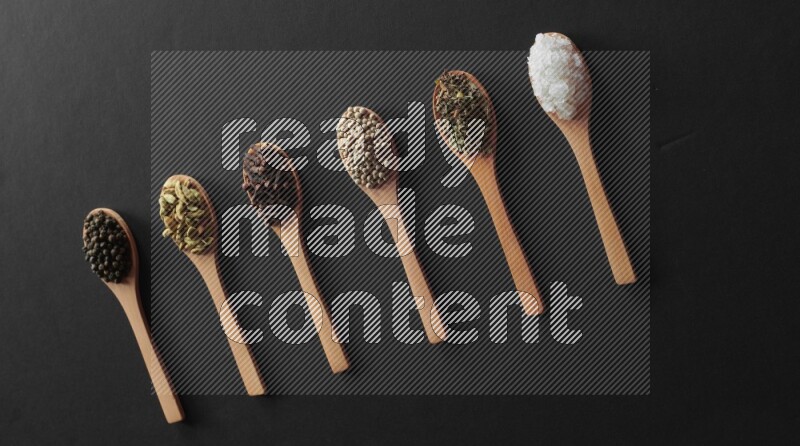 wooden spoons filled with white peppers, cloves, cardamom, salt, black peppers and basil on black flooring
