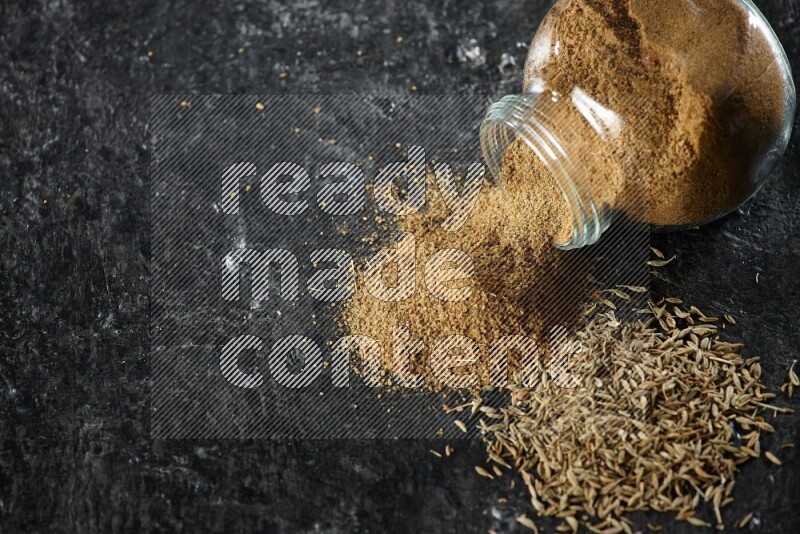 A flipped glass spice jar full of cumin powder with spilled powder and cumin seeds on a textured black flooring