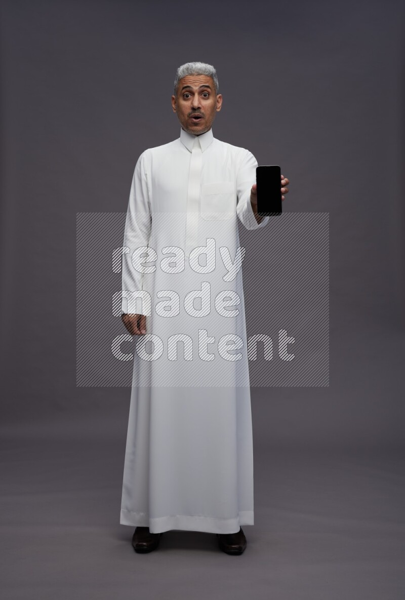 Saudi man wearing thob standing showing phone to camera on gray background