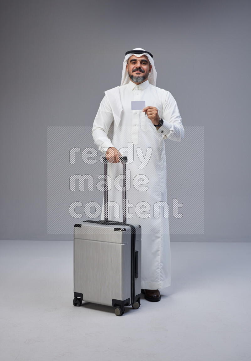 Saudi man wearing Thob and white Shomag standing holding Travel bag and ATM card on Gray background