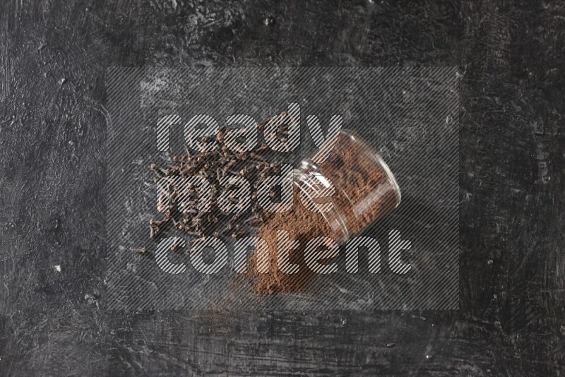 A flipped glass jar full of cloves powder with cloves spread on a textured black flooring