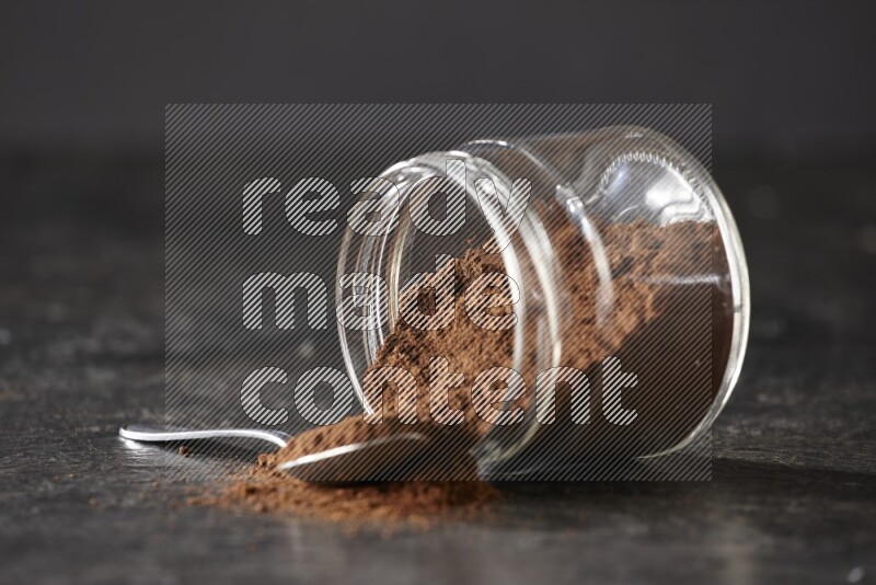 A flipped glass jar and metal spoon full of cloves powder with cloves spread on a textured black flooring