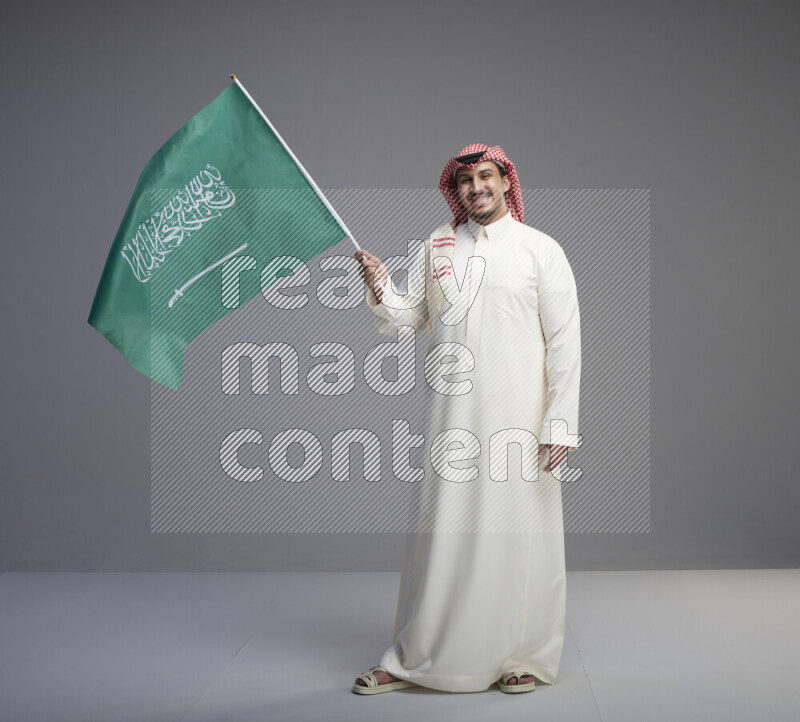 A Saudi man standing wearing thob and red shomag raising big Saudi flag on gray background