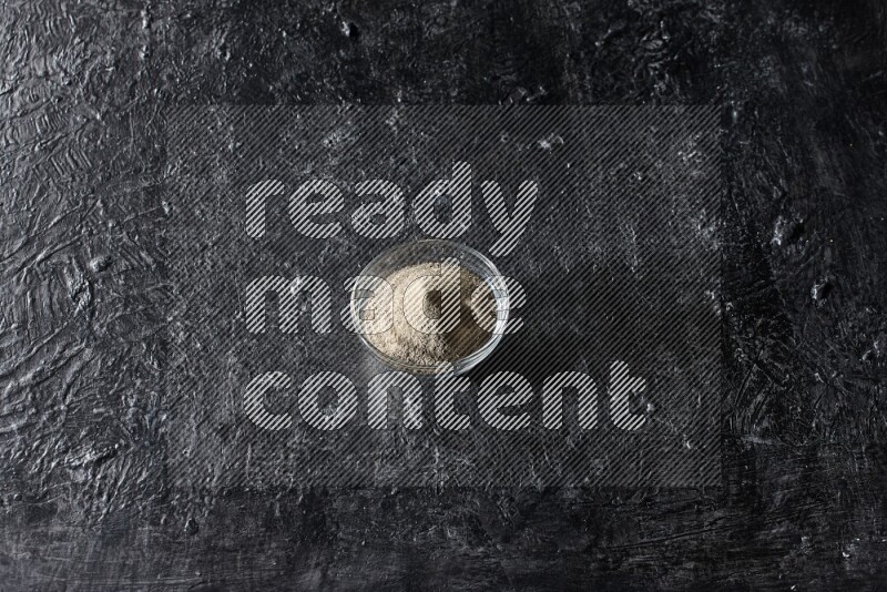 A glass bowl full of white pepper powder on textured black flooring