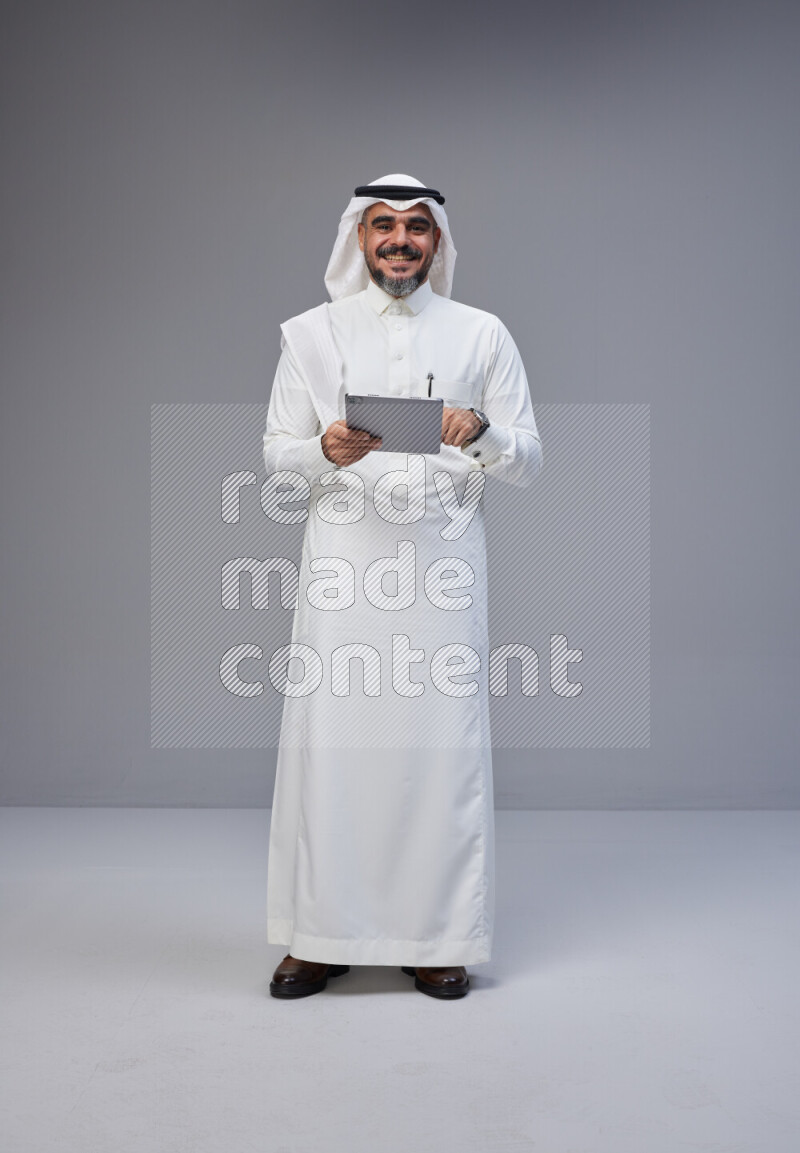 Saudi man Wearing Thob and white Shomag standing working on tablet on Gray background