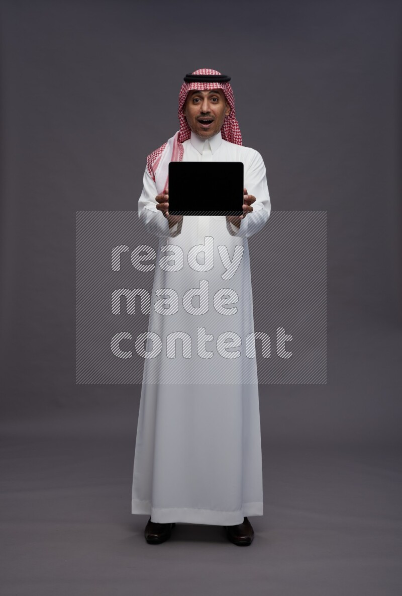 Saudi man wearing thob and shomag standing showing tablet to camera on gray background
