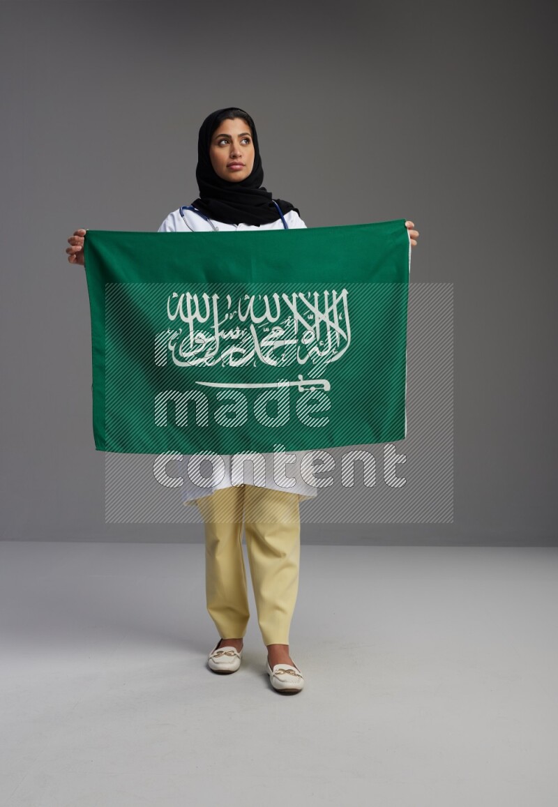 Saudi woman wearing lab coat with stethoscope standing holding Saudi flag on Gray background