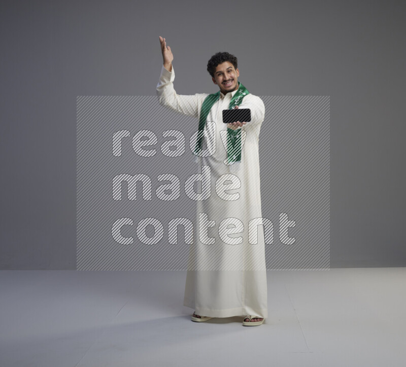 A Saudi man standing wearing thob and Saudi flag scarf showing phone to camera on gray background