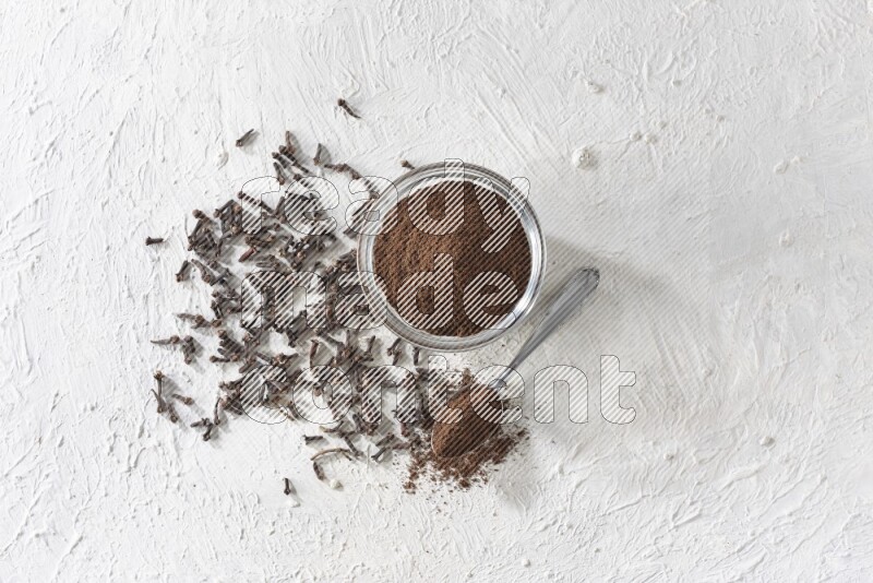 A glass bowl and a metal spoon full of cloves powder with cloves grains spread on a textured white flooring