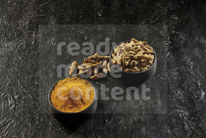 2 pottery black bowls, one full of turmeric powder and the other full of dried turmeric whole fingers on a textured black flooring