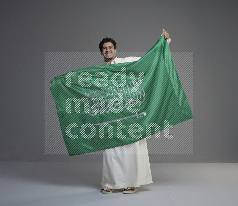 A Saudi man standing wearing thob holding big Saudi flag on gray background