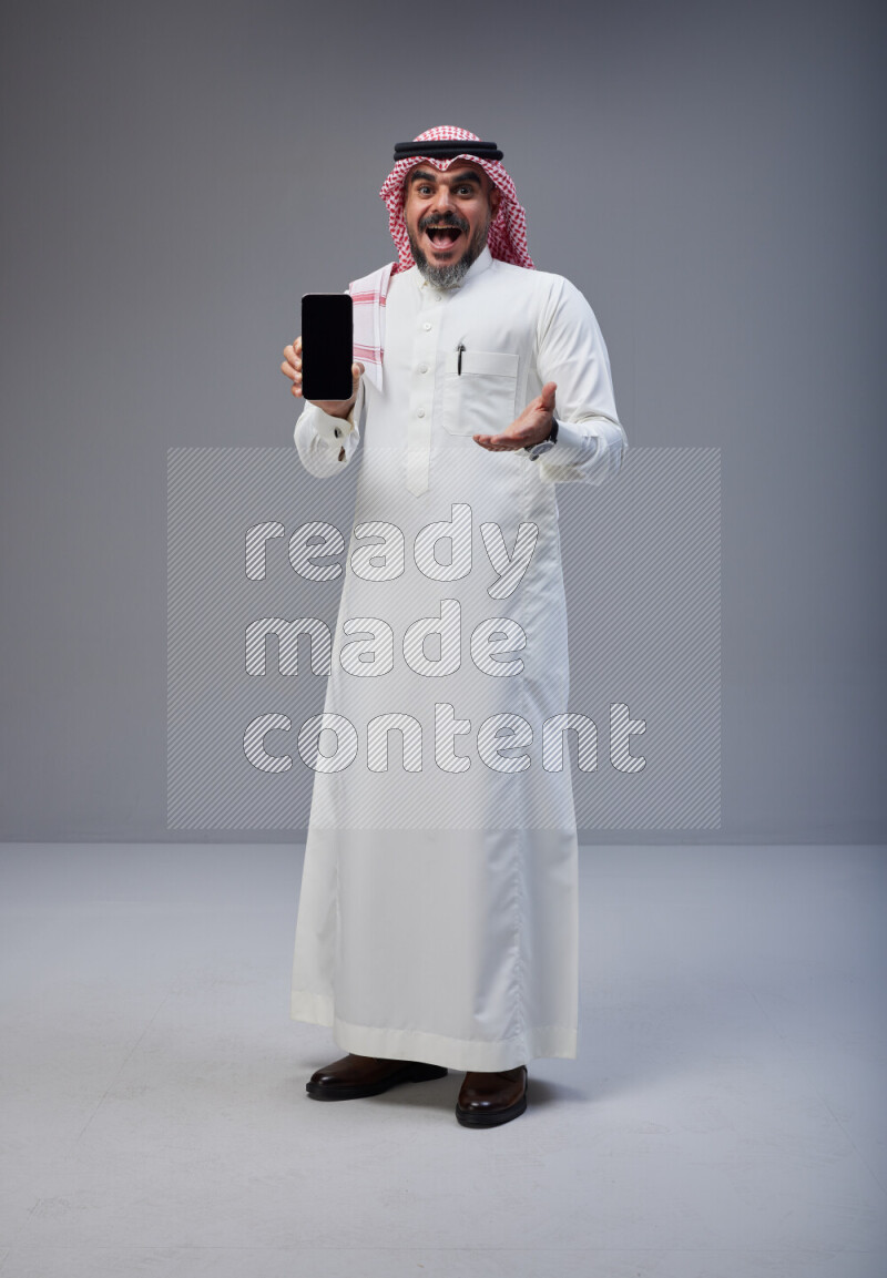 Saudi man Wearing Thob and red Shomag standing showing phone to camera on Gray background