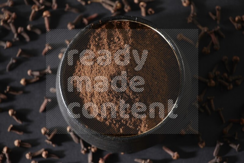 A black pottery bowl full of cloves powder and cloves whole grains on a black flooring