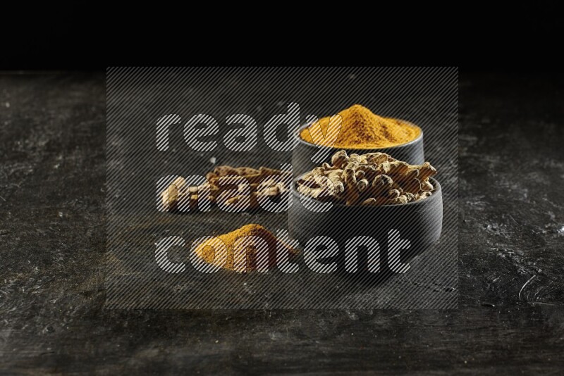 2 pottery black bowls, one full of turmeric powder and the other full of dried turmeric whole fingers on a textured black flooring