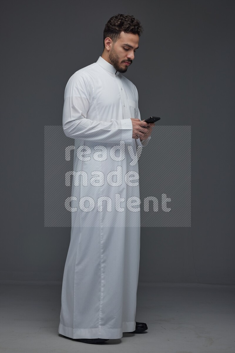 A Saudi man wearing Thobe standing and using his phone eye level on a gray background