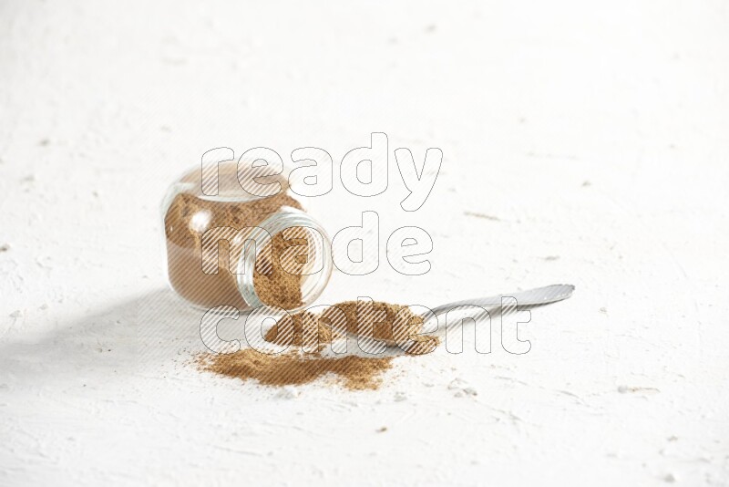Flipped herbs glass jar full of cinnamon powder with a metal spoon full of powder on a textured white background