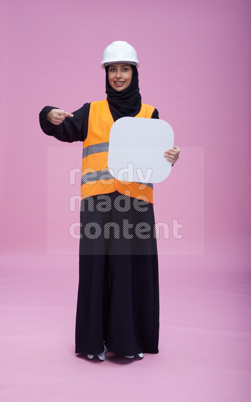 Saudi woman wearing Abaya with engineer vest and helmet standing holding social media sign on pink background
