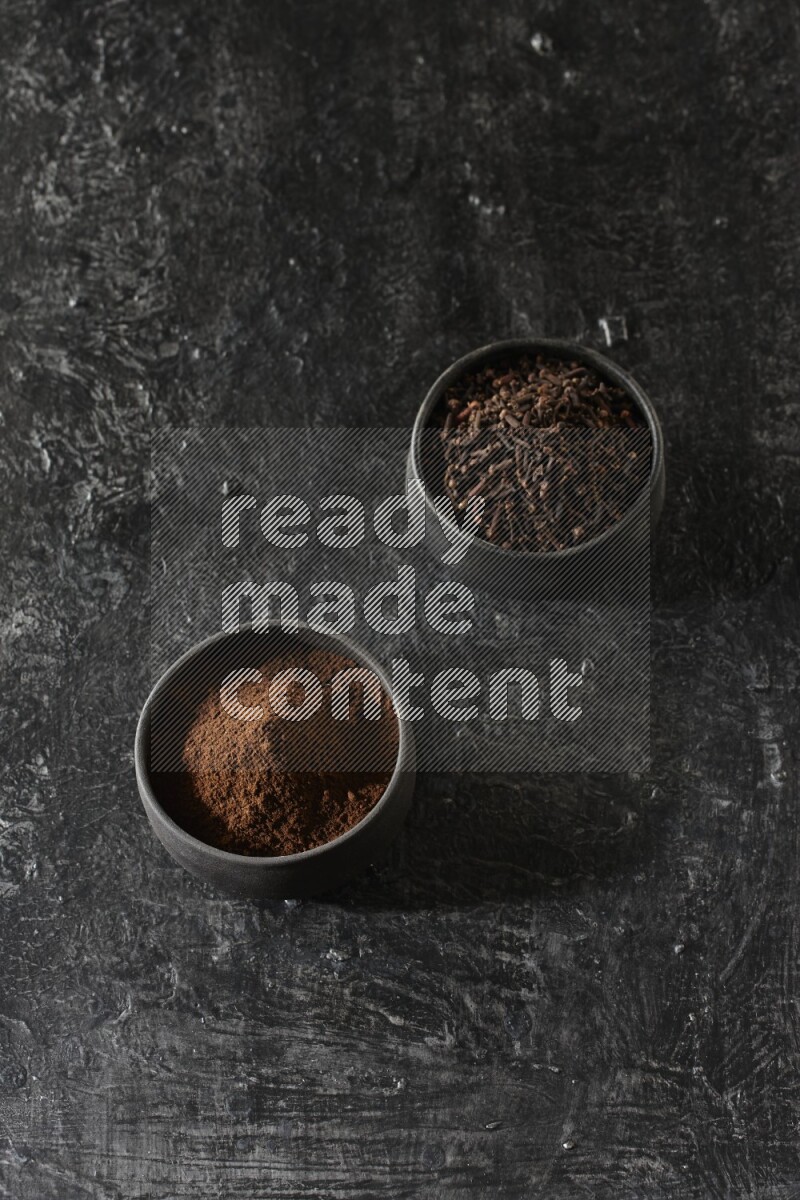 2 Black pottery bowls full of cloves and the other full of cloves powder on textured black flooring
