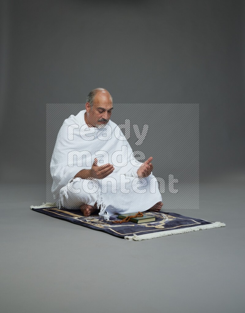 A man wearing Ehram sitting on prayer mat dua'a on gray background