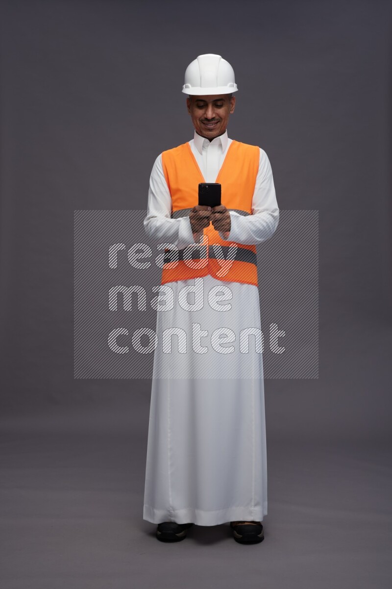 Saudi man wearing thob with engineer vest standing texting on phone on gray background