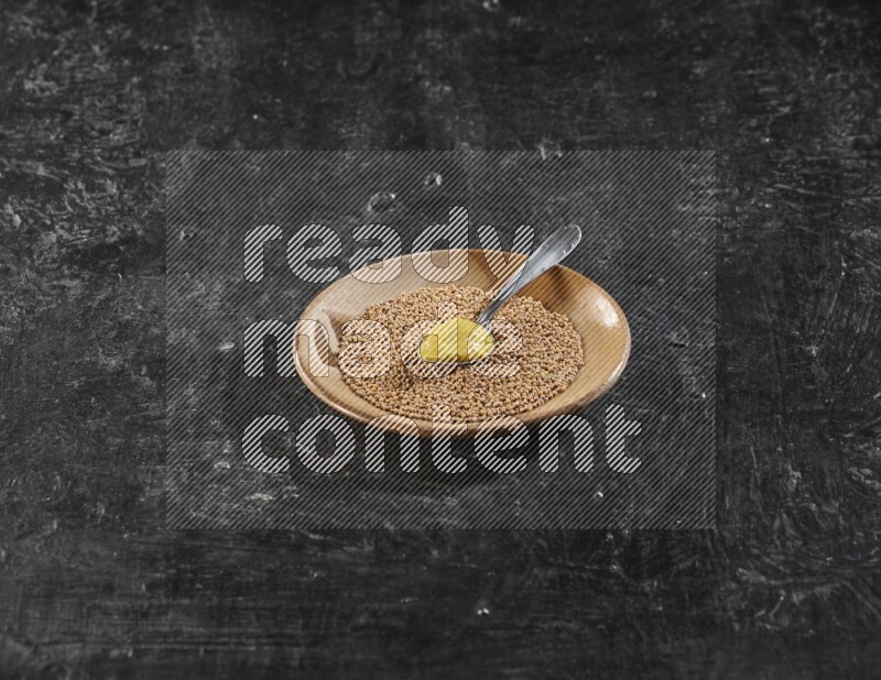 A beige pottery plate filled with mustard seeds with a metal spoon full of mustard paste in it on a textured black flooring