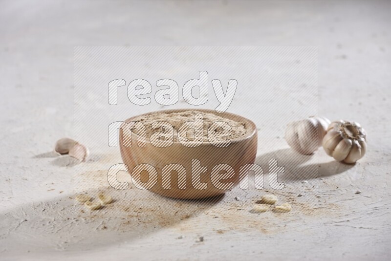 A wooden bowl full of garlic powder and beside it garlic cloves on a textured white flooring in different angles