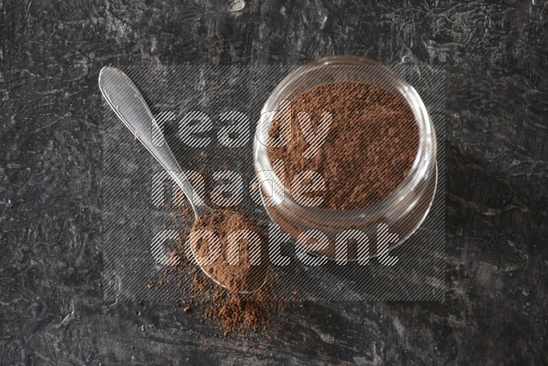 A glass jar full of cloves powder with a metal spoon on a textured black flooring