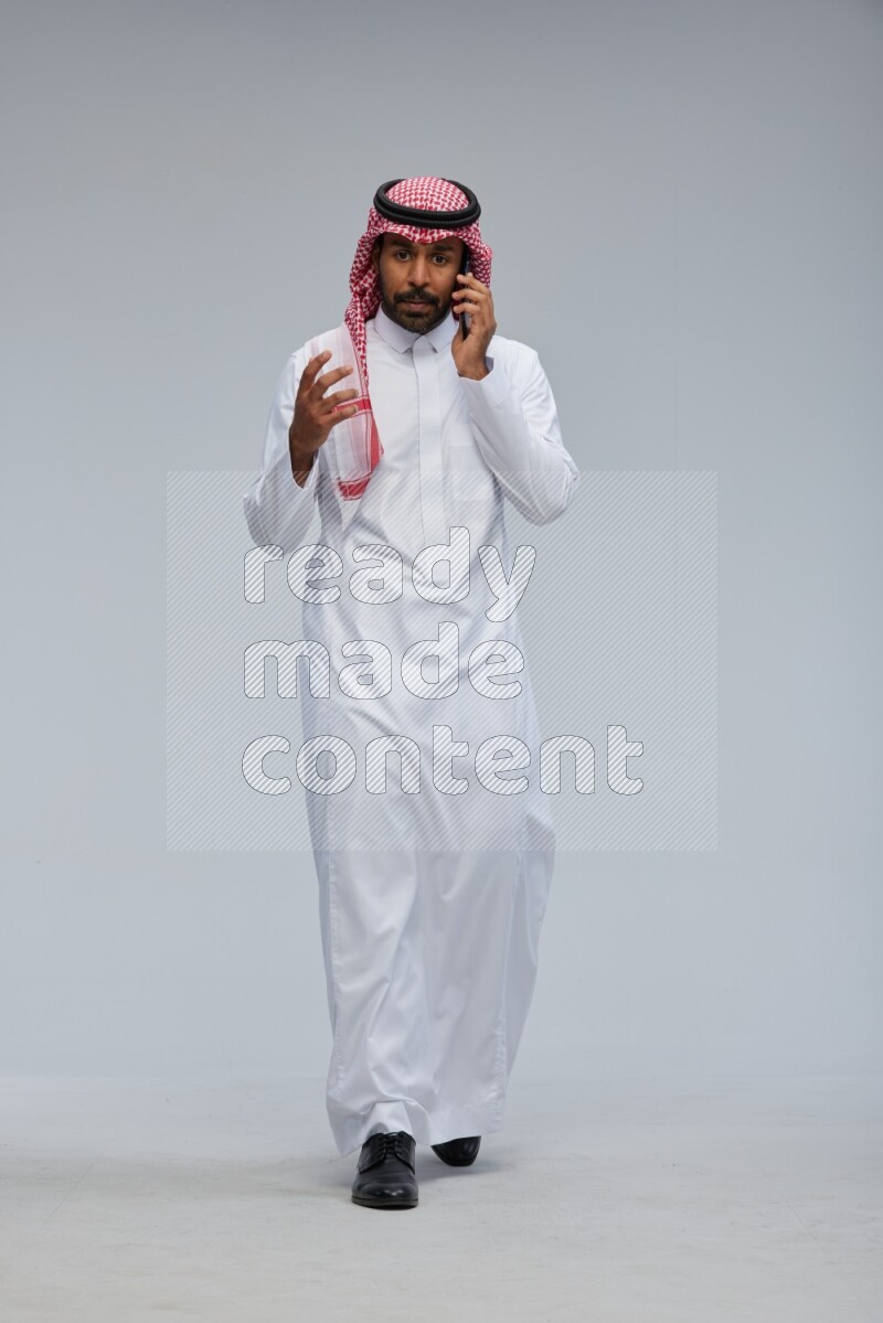 Saudi man Wearing Thob and shomag standing talking on phone on Gray background