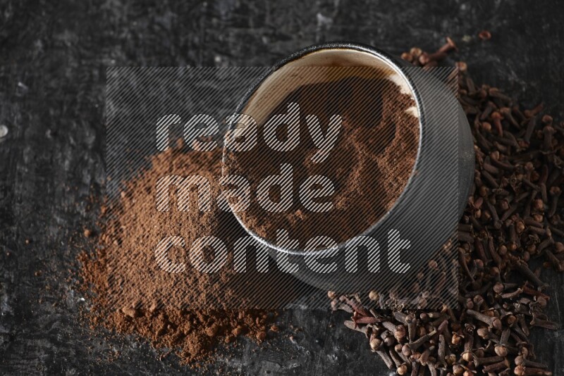 وعاء فخاري أسود ممتلئ ببودرة القرنفل والبودرة تناثرت منه مع الحبوب على خلفية سوداء