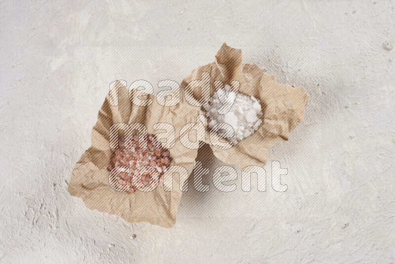 2 crumpled pieces of paper full of pink himalayan salt and white salt on white background