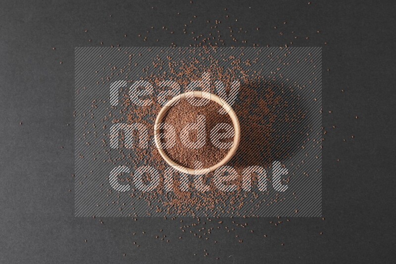 A wooden bowl full of garden cress seeds surrounded by seeds on a black flooring