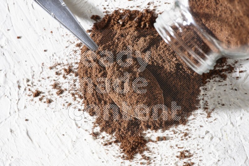 A flipped glass spice jar and a metal spoon full of cloves powder and powder came out of the jar on textured white flooring