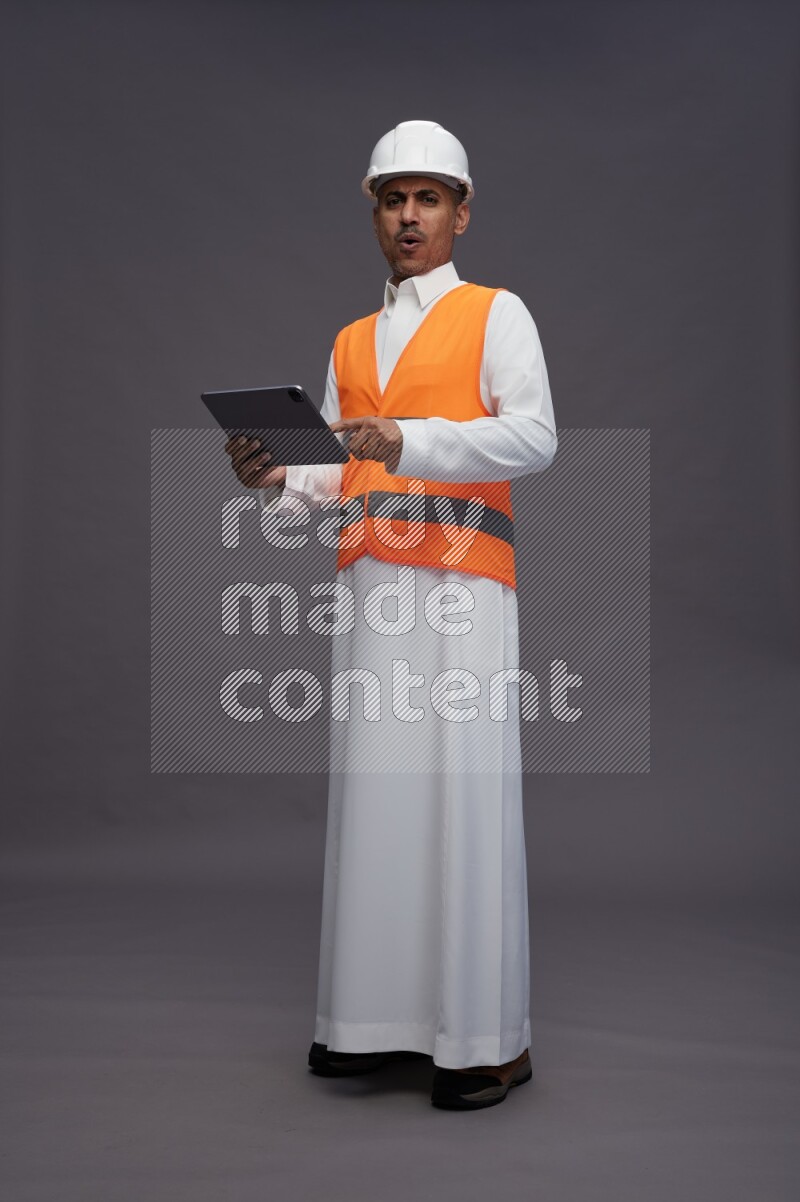Saudi man wearing thob with engineer vest standing working on tablet on gray background