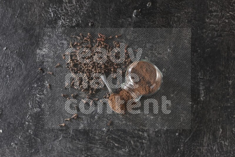 A flipped glass spice jar and a metal spoon full of cloves powder with cloves spread on textured black flooring