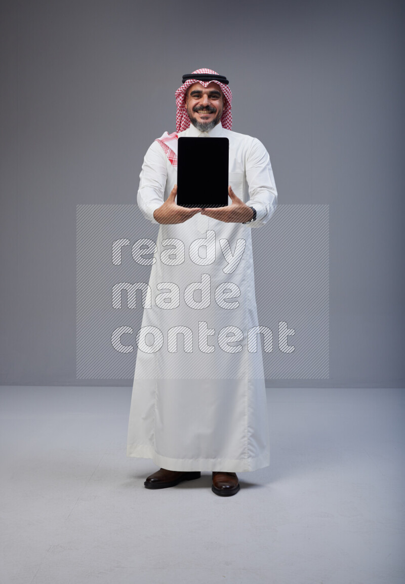 Saudi man Wearing Thob and red Shomag standing showing tablet to camera on Gray background