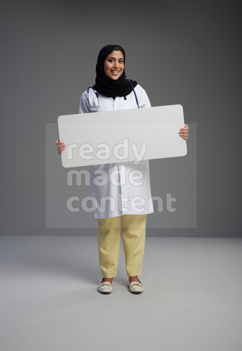 Saudi woman wearing lab coat with stethoscope standing holding board on Gray background